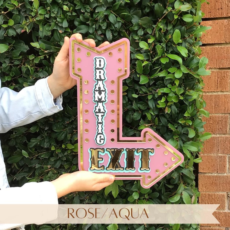 Features rose pink acrylic "Dramatic Exit" arrow sign with gold mirror accents. Motivational, empowering decor for eclectic, industrial, modern, bright, and boho spaces. Perfect for bedrooms, salons, craft rooms, cafes, and gift for confident women. Customizable arrow direction & color. Bright, bold, confidence-boosting home decor, dopamine decor art, quirky wall hanging, vintage marquee wall art, inspiring wall decor, affirmation sign, motivational decor, eclectic home decoration.