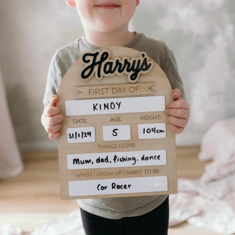 Op de afbeelding: Een houten bord met de naam "Harry's" geschreven in zwarte cursieve letters. Het bord is versierd met een banner waarop staat "Eerste dag van: KINOY". Het bord bevat ook een datum, een leeftijd, een lengte en dingen die het kind leuk vindt. De datum is 31/1/24, de leeftijd is 5, de lengte is 104 cm en de dingen die het kind leuk vindt zijn "Mama, papa, vissen, dansen". Het bord bevat ook een sectie voor "Wanneer ik groot ben, wil ik zijn:" en het kind heeft "Autocoureur" geschreven.