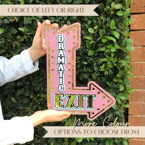 May include: A pink arrow-shaped sign with gold trim and gold circular accents. The sign features the text "DRAMATIC" vertically and "EXIT" horizontally in a bold, dark font. The text "CHOICE OF LEFT OR RIGHT" and "MORE COLOUR OPTIONS TO CHOOSE FROM" appear at the top and bottom.