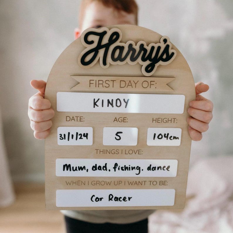 Op de afbeelding: Een houten bord met de naam "Harry's" geschreven in zwarte cursieve letters. Het bord is versierd met een banner met de tekst "Eerste dag van: Kinoy". Het bord bevat ook de datum "31/1/24", de leeftijd "5", de lengte "104 cm", dingen die de persoon leuk vindt en wat ze wil worden als ze groot is.