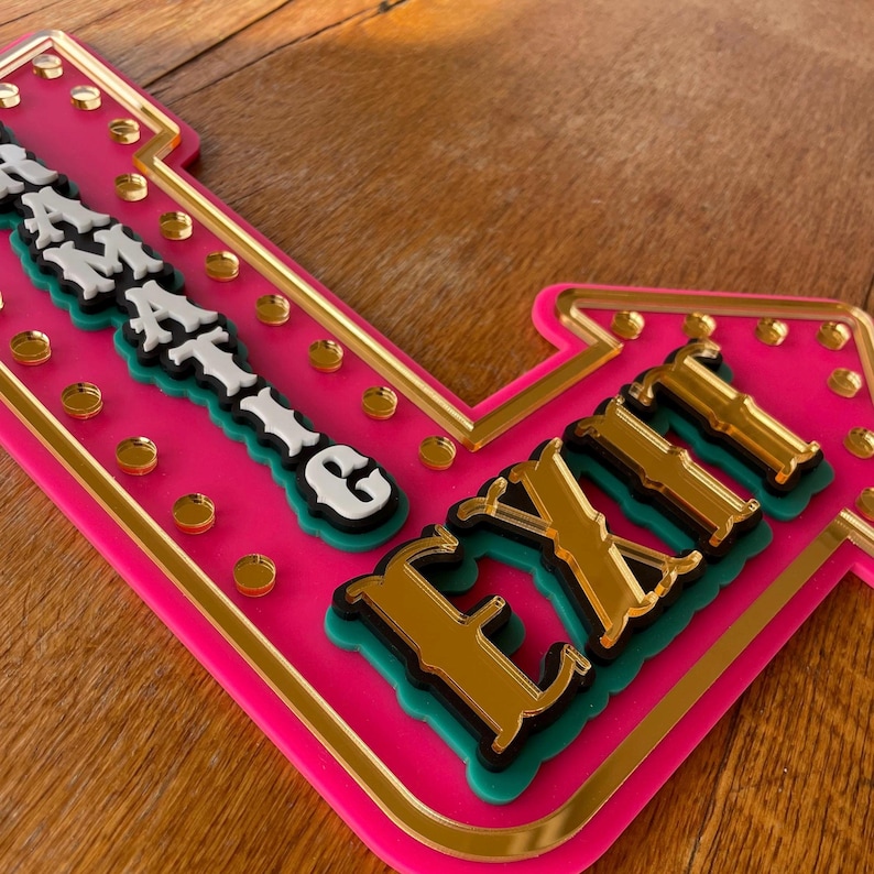 May include: A vibrant pink and gold sign with the word "EXIT" in gold mirrored letters. The sign also features the word "AMAZING" in white and black letters. The sign is decorated with gold-colored circular accents.