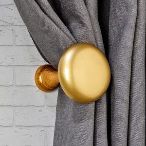 Può includere: Un fermatenda dorato con un design rotondo e piatto. Il fermatenda tiene una tenda grigia. La tenda ha un aspetto strutturato. Il fermatenda è montato su un muro di mattoni bianchi.