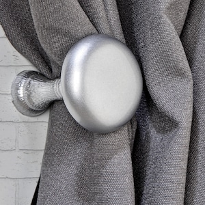 May include: A silver curtain tieback with a round, smooth head and a decorative base, holding a gray textured curtain. The tieback is mounted on a white brick wall, creating a contrast of textures and colors.