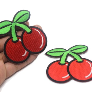 May include: Two red cherries with green stems and leaves. The cherries are embroidered on a white fabric.
