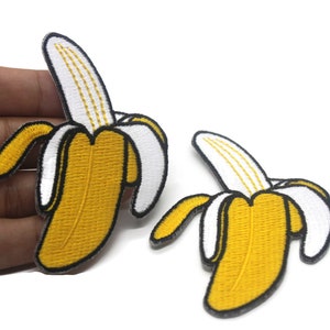 Puede incluir: Dos parches bordados de plátano amarillo con contornos blancos. Los plátanos son curvos y tienen un contorno negro.