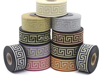 May include: A stack of spools of ribbon with a Greek key pattern in various colors, including black, white, gold, silver, red, and purple.
