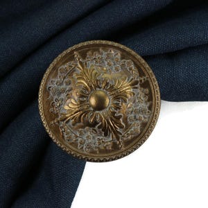 Peut inclure: Un embrasse de rideau en métal doré avec un motif orné comprenant un motif floral central. L'embrasse est sur un fond de tissu bleu foncé.