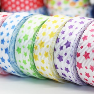 May include: A set of six rolls of white ribbon with different colored stars printed on them. The ribbon colors are red, blue, green, yellow, purple, and pink.