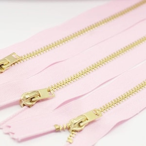 May include: Four pink zipper tapes with gold metal teeth and sliders. The zippers are laid out in a row, with the sliders facing the viewer.