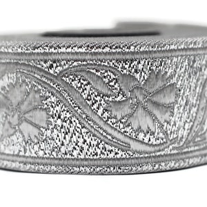 May include: A silver-colored, cylindrical object with an intricate floral pattern. The design is embossed around the circumference, creating a textured appearance. The object is set against a white background.