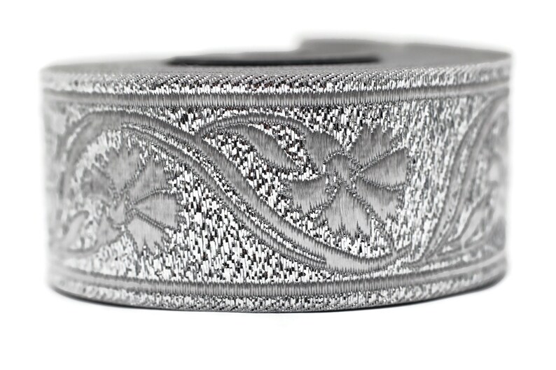 May include: A silver-colored, cylindrical object with an intricate floral pattern. The design is embossed around the circumference, creating a textured appearance. The object is set against a white background.