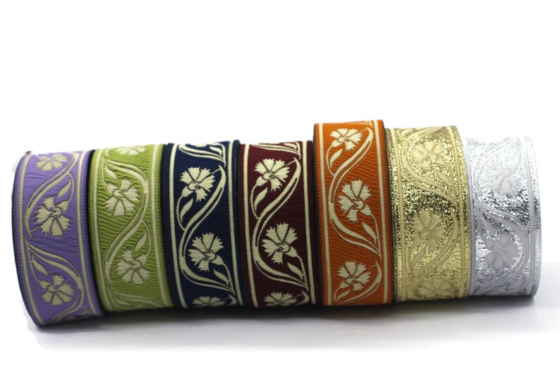 May include: A collection of decorative ribbons in various colors, including lavender, green, navy, burgundy, orange, gold, and silver. Each ribbon features a floral design with a repeating pattern. The ribbons are rolled up, showcasing their intricate details.