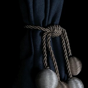 Pode incluir: Cortina de tecido azul escuro com um amarrador de corda marrom e três borlas decorativas. Duas das borlas são brancas e uma é marrom.