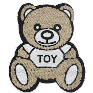 Op de afbeelding: Een bruine teddybeer met een wit shirt waarop "TOY" staat. De beer zit met gekruiste benen.