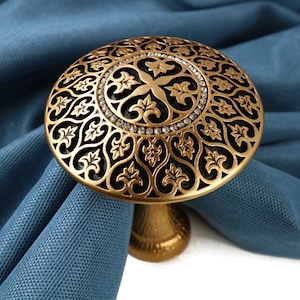 May include: Gold curtain tieback with a black and gold ornate design. The tieback has a round top with a decorative pattern and a small base.