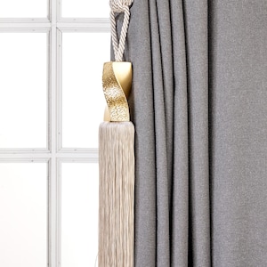May include: A gold-toned tassel tieback with a twisted design and a long, cream-coloured fringe. The tieback is attached to a cream-coloured rope.
