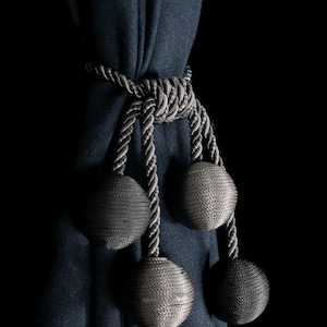 Pode incluir: Uma cortina azul escura com um amarrador de corda prateado e cinza. O amarrador tem quatro borlas redondas, prateadas e cinzas.