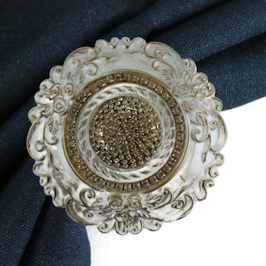 Könnte beinhalten: Ein dekorativer Vorhangbinder mit weißer und goldener Oberfläche. Der Binder hat ein rundes Design mit filigranen Blumendetails und einem zentralen Stück aus funkelnden Strasssteinen.