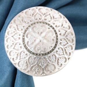 Puede incluir: Un sujeta cortinas de plata con un diseño intrincado y ornamentado. El sujeta cortinas tiene una forma circular con un patrón floral en relieve y un círculo central de diamantes de imitación.