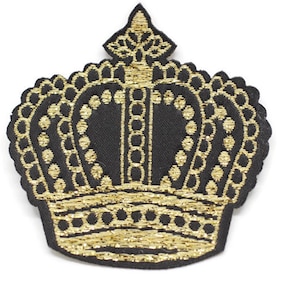 Puede incluir: Un parche de corona bordado en oro sobre un fondo de tela negro. La corona tiene una punta puntiaguda y un diseño detallado.