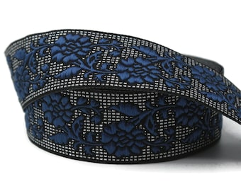 Puede incluir: Cinta decorativa enrollada con un diseño floral. La cinta tiene un patrón floral bordado en azul oscuro sobre un fondo plateado y negro. La cinta se utiliza probablemente para manualidades o adornos.
