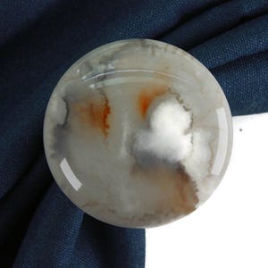 Puede incluir: Un sujeta cortinas redondo de ágata blanca con remolinos marrones y grises. El sujeta cortinas está sobre un fondo de tela azul oscuro.