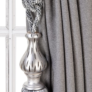 May include: A silver curtain tieback with a tassel and a decorative, ornate, silver,  bulbous finial. The tieback is holding back a gray curtain.