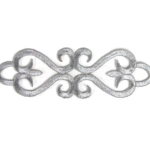 Puede incluir: Adorno bordado en plata con un diseño decorativo y ornamentado. El adorno presenta un patrón repetitivo de espirales y bucles.