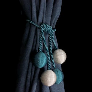Pode incluir: Um amarrador de cortina azul-turquesa com três borlas redondas. Duas das borlas são azul-turquesa e uma é de cor creme claro.