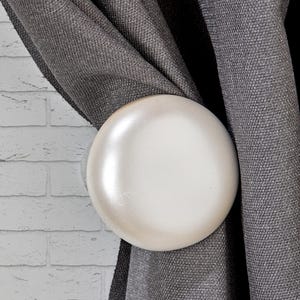 May include: A close-up of a white, round curtain tieback. The tieback is holding a section of gray fabric curtains. The background includes a white brick wall and more gray fabric.