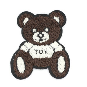 Op de afbeelding: Bruine teddybeer pluche patch met witte accenten en het woord "TOI" op de borst.