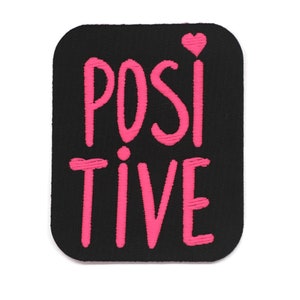 Puede incluir: Parche de tela negro con texto bordado en rosa que dice "POSITIVE" con un pequeño corazón rosa sobre la "I".