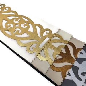 Può includere: Un set di campioni di tessuto stratificati con un intaglio in filigrana metallica dorata sopra pannelli di tessuto bianco, beige e grigio scuro. Il pezzo dorato presenta un motivo a volute complesso e simmetrico con una finitura liscia.