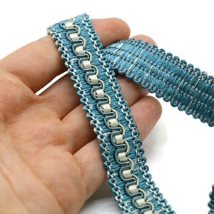 May include: Close-up of a decorative trim in shades of teal and off-white. The trim features a woven texture with a repeating pattern of loops and a scalloped edge. The trim is likely used for embellishing clothing or home decor items.