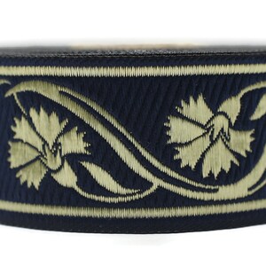 May include: A navy blue hat with an embroidered floral design in a light gold color. The hat features a black band at the top and bottom, and the floral pattern wraps around the hat's circumference.