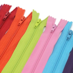 Puede incluir: Un conjunto de ocho cierres de cremallera en varios colores, incluyendo morado, azul, rosa, verde, naranja y rojo. Las cremalleras están dispuestas en una fila, con la cremallera morada a la derecha y la cremallera roja a la izquierda.