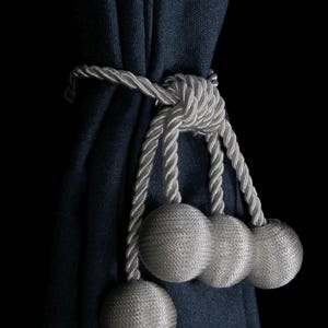 Pode incluir: Uma cortina azul escura com um amarrador de corda branco com três pompons redondos brancos.