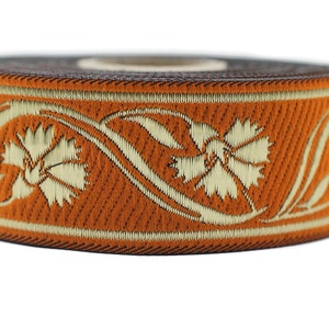 May include: A roll of orange ribbon with a floral design in cream-colored thread. The ribbon features a repeating pattern of stylized flowers and vines, with a dark brown edge.