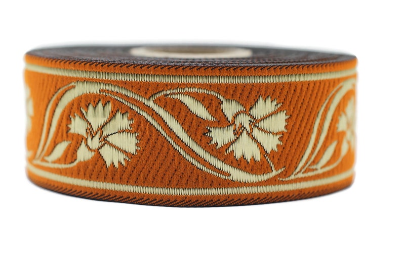 May include: A roll of orange ribbon with a floral design in cream-colored thread. The ribbon features a repeating pattern of stylized flowers and vines, with a dark brown edge.