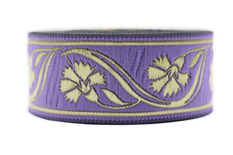 May include: A roll of decorative ribbon featuring a floral design. The ribbon has a lavender background with gold-colored embroidered flowers and accents. The edges are dark purple.