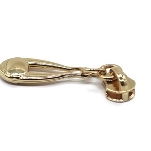 May include: Gold zipper pull with a decorative oval-shaped top and a chain link connecting to the zipper slider.