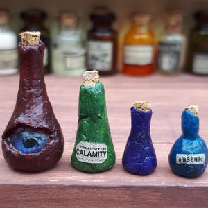 Puede incluir: Cuatro botellas de vidrio en miniatura con tapones de corcho. Las botellas están llenas de líquidos de diferentes colores. Las botellas están etiquetadas con las palabras "Calamity" y "Arsenic".