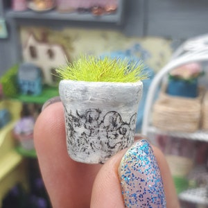 Puede incluir: Una maceta de cerámica blanca en miniatura con un diseño en blanco y negro, llena de hierba artificial verde. La maceta está siendo sostenida por una mano con esmalte de uñas azul con purpurina.