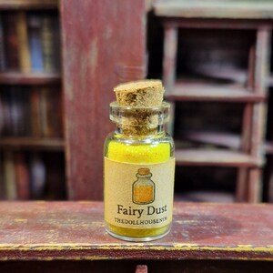 Puede incluir: Una pequeña botella de vidrio llena de polvo amarillo, etiquetada como "Fairy Dust". La botella tiene un tapón de corcho y una etiqueta de papel con un dibujo de una botella. El texto "thedollhousentm" está debajo de las palabras "Fairy Dust".