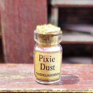 Puede incluir: Una pequeña botella de vidrio etiquetada como "Pixie Dust" con un tapón de corcho. La botella contiene contenido rosa y dorado. La etiqueta es de color beige claro con texto marrón oscuro. El fondo es una estructura de madera borrosa.