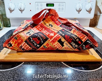 May include: A red, black, and tan BBQ-themed food carrier with a handle, resting on a wooden cutting board. The fabric features text and graphics related to barbecue, including the words "BBQ," "Grill," and "Smokehouse."
