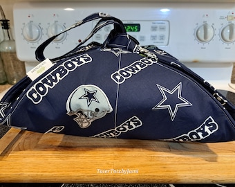 May include: A blue and white fabric food warmer with a Dallas Cowboys theme. The warmer has a star and the words "Cowboys" printed on it. The warmer is sitting on a wooden cutting board.