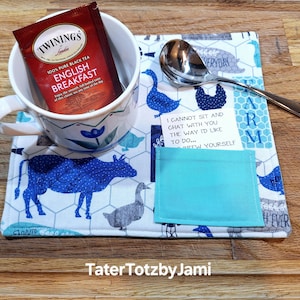 May include: A white mug with a blue floral design holds a tea bag. The mug sits on a farm-themed placemat with blue cow, goose, and chicken illustrations. A silver spoon rests on the placemat, which has the text "I cannot sit and chat with you the way I'd like to do... Brew yourself".
