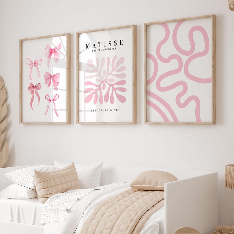 K&ouml;nnte beinhalten: Drei gerahmte Kunstdrucke mit rosafarbenen abstrakten Designs. Die Drucke h&auml;ngen an einer wei&szlig;en Wand &uuml;ber einem Bett mit einer beigefarbenen Decke und Kissen.