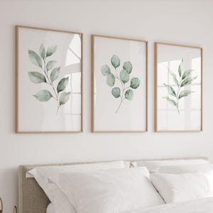 Puede incluir: Tres impresiones botánicas enmarcadas con ilustraciones de acuarela de hojas y ramas verdes. Las impresiones tienen marcos de madera marrón claro y se exhiben en una pared blanca sobre una cama con ropa de cama blanca. La obra de arte añade un toque de naturaleza.